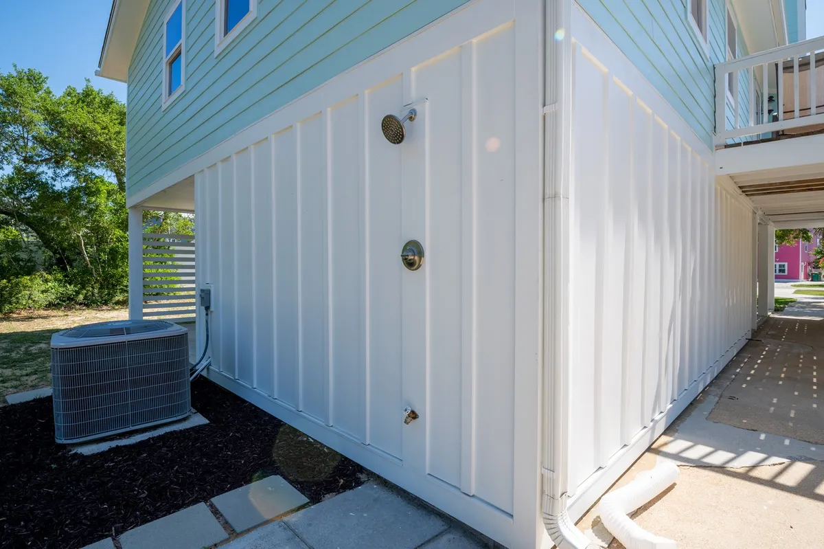 A fresh and clean outdoor shower area is positioned beside the house, with visible siding details and air conditioning unit. The shower offers a practical spot to rinse off after beach outings, connecting directly to the outdoor adventure theme.