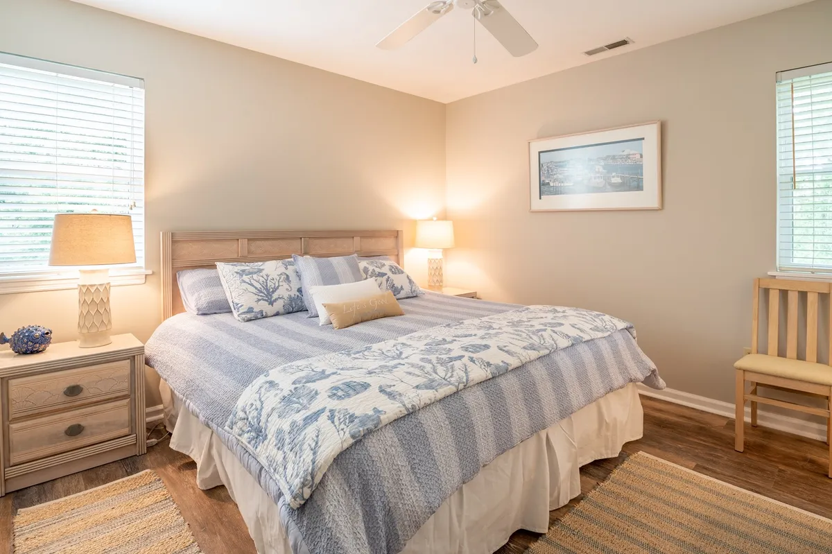 A serene bedroom featuring a bed with blue and white bedding, a nightstand with a lamp, and a cozy chair by the window. Framed artwork adorns the wall, and ample natural light streams in through the window, creating a restful environment.