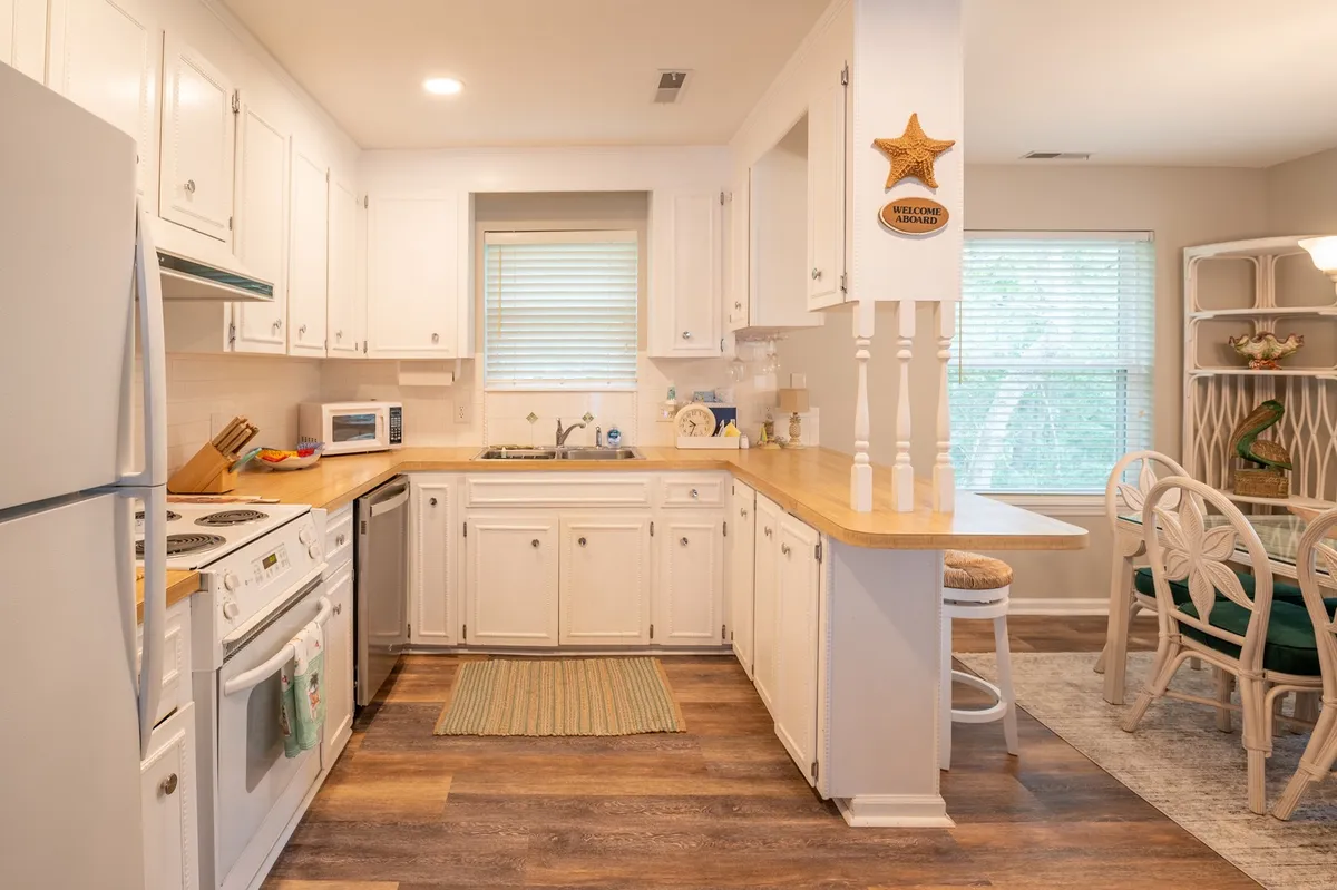 A welcoming view of a kitchen and dining area, featuring a large kitchen island, wooden countertops, and coastal-themed decorations like a starfish. Windows with blinds let in natural light, and the layout is open and inviting for group gatherings.