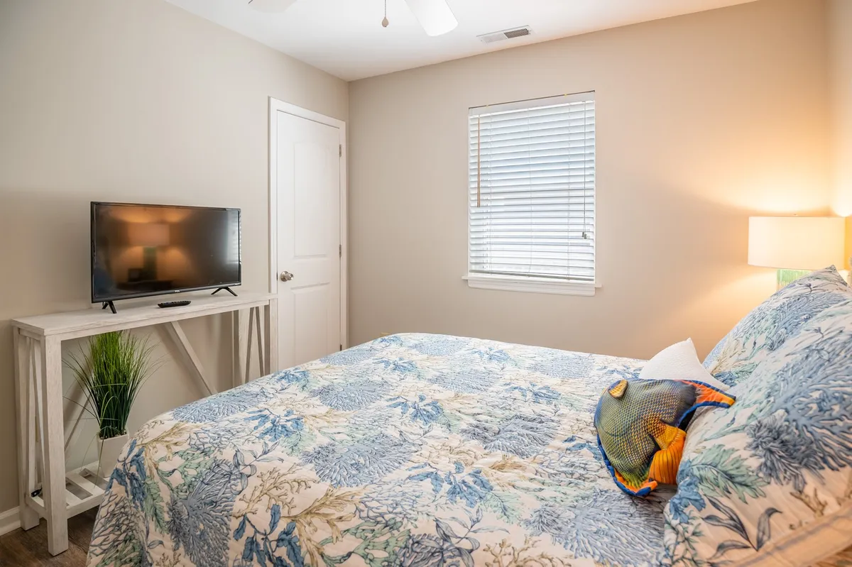 A cozy guest bedroom with floral bedding, a television on a stand, a lamp casting warm light, and a small potted plant. Sunlight filters through the window, enhancing the calm ambiance, while the TV and seating make it ideal for winding down.