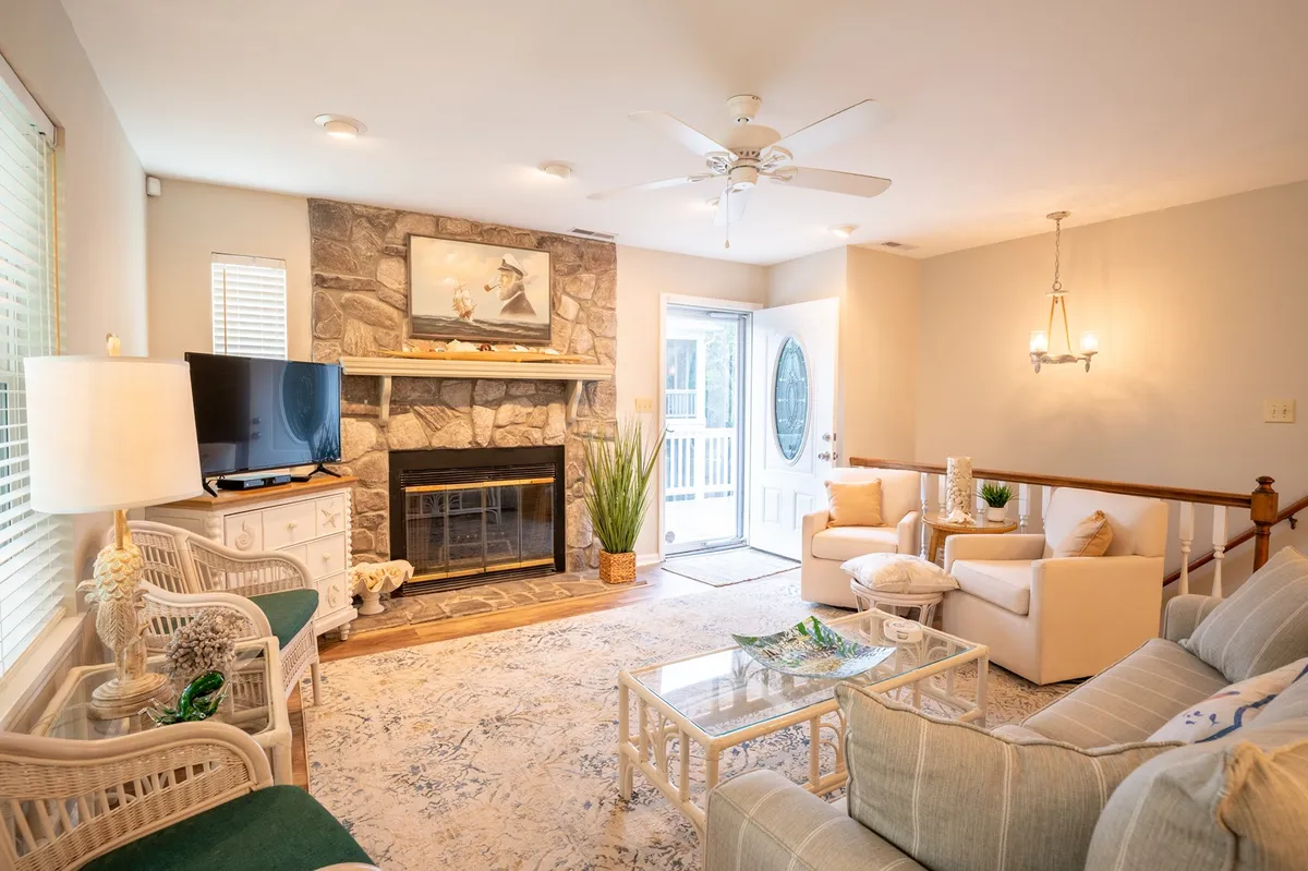 This image showcases a spacious living room with a stone fireplace, large windows allowing in natural light, comfortable seating, and a television mounted above the fireplace. The room exudes a cozy and inviting atmosphere, perfect for relaxing or gathering with family and friends.