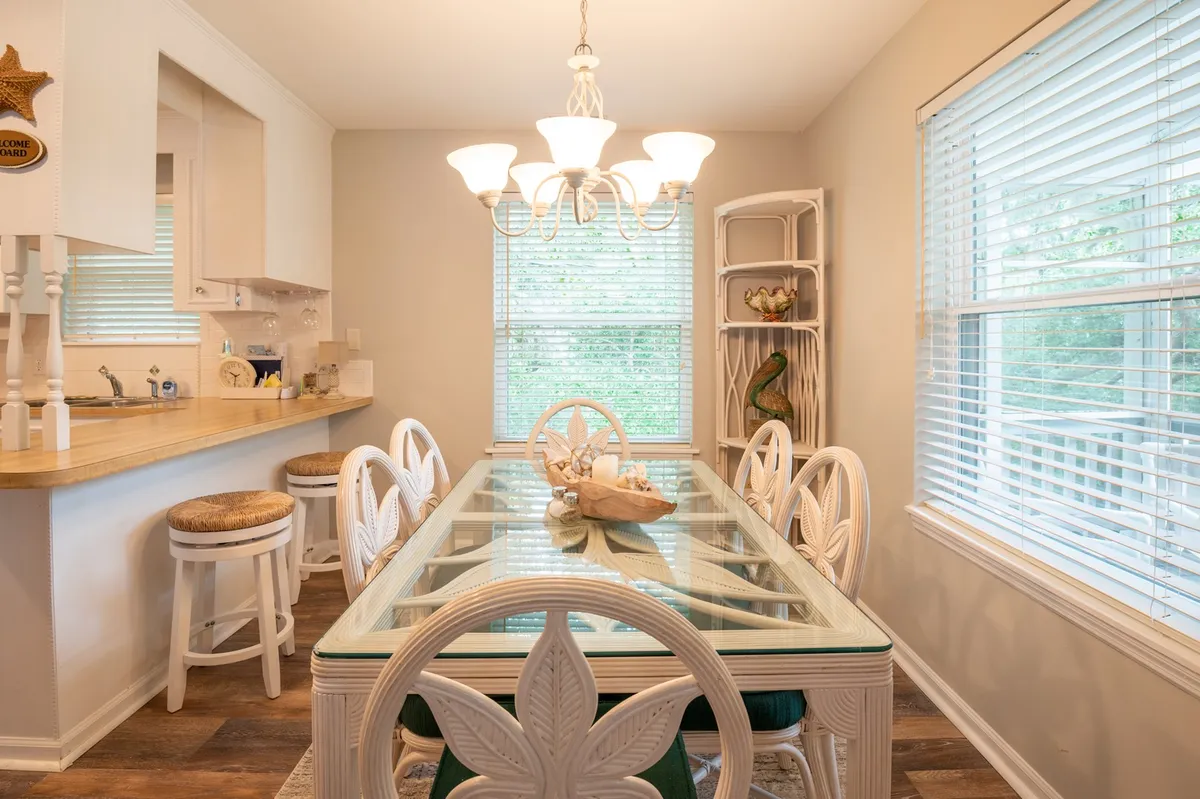 A welcoming dining area with a central dining table, elegant chandelier, large window streaming in natural light, and a decorative shelf. The space is set up for family meals, entertaining guests, or casual gatherings.
