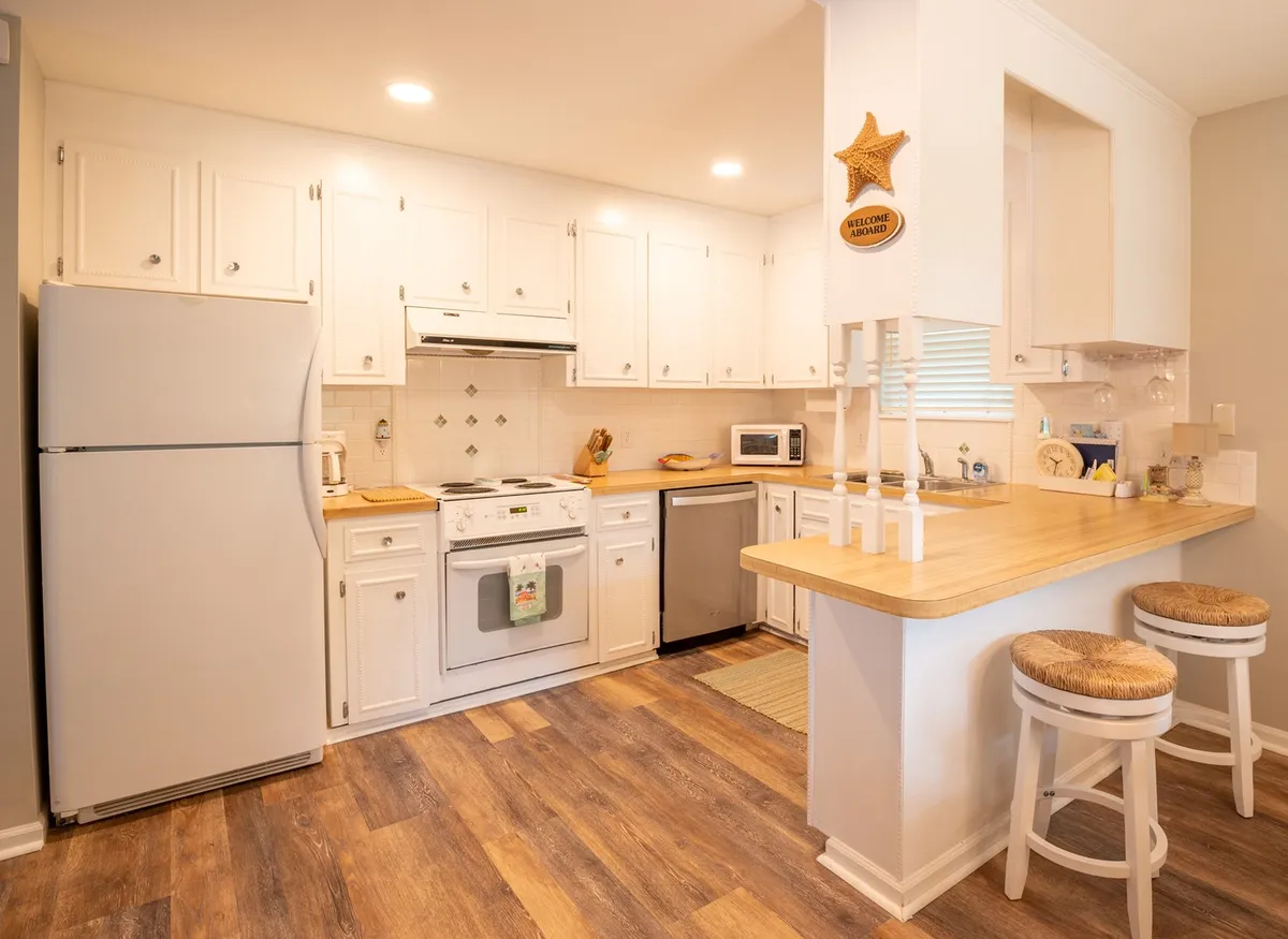 A view of a bright, fully equipped kitchen featuring white cabinetry, a wooden countertop, stainless steel appliances, and an inviting bar top with stools—ideal for preparing meals or enjoying quick bites together.