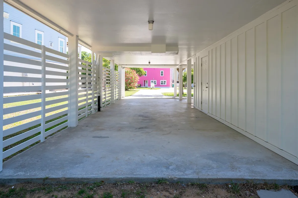 This image shows an open carport with white slatted walls and a pink house in the background. The carport offers covered parking space, while the driveway can accommodate multiple vehicles. The area is shaded and welcoming, creating a practical and inviting first impression.