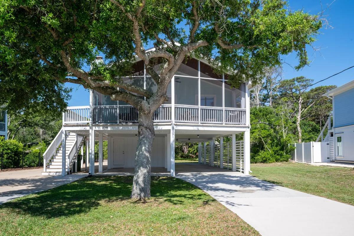 A broad view of the exterior, with a large tree shading the driveway and the house’s welcoming porch in the background. The driveway suggests ample parking, while the porch and greenery create an inviting, peaceful atmosphere ideal for family gatherings and relaxed outdoor living.