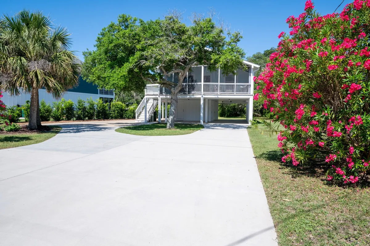 This image displays the exterior of The Holland House set amidst flowering bushes, a prominent palm tree, and a welcoming porch area. The scene exudes a relaxed coastal vibe, with natural greenery framing the house and hints of sunset light indicating the tranquil end of a beach-filled day.